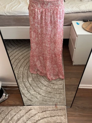 Långkjol med rosa paisleymönster - Säljer en flowy långkjol i rosa och vitt med paisleyinspirerat mönster. Kjolen har resår i midjan och är fodrad med ett vitt innerfoder. Perfekt för dig som gillar bohemiska vibes och vill ha något bekvämt men ändå snyggt.