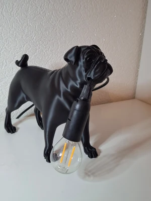 Bordslampa mops  - Unik bordslampa formad som en Mops i svart matt material. Lampan har en glödlampa som hänger från hundens mun och en sladd baktill. Perfekt som cool inredningsdetalj för dig som gillar hundar och vill ha något annorlunda i rummet.