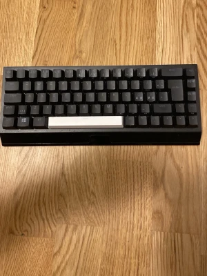 Razer Wireless Mechanical Keyboard - Säljer ett svart kompakt mekaniskt tangentbord med vita tangenter för mellanslag. Tangentbordet har en minimalistisk design och passar perfekt för dig som vill ha ett smidigt och stilrent tillbehör till datorn. Materialet är plast och layouten är tenkeyless.