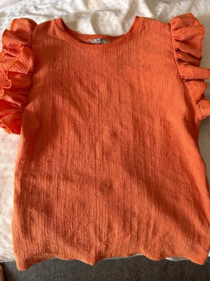 Orange topp med volangärmar - Säljer en snygg orange topp med korta volangärmar. Toppen har en strukturerad yta och rund halsringning. Perfekt för dig som vill sticka ut med färg och coola detaljer. Passar till både jeans och kjol. Köpt utomlands så är osäker på märket