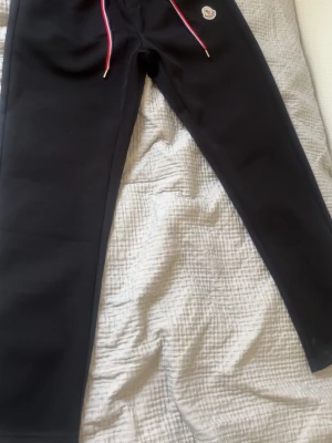 Svarta mjukisbyxor med logga - Säljer ett par svarta mjukisbyxor med dragsko i midjan och detaljer i rött och vitt på snörena. Byxorna har en liten logga framtill och en ficka bak med glansig detalj. Perfekta för chill dagar och riktigt sköna att ha på sig.