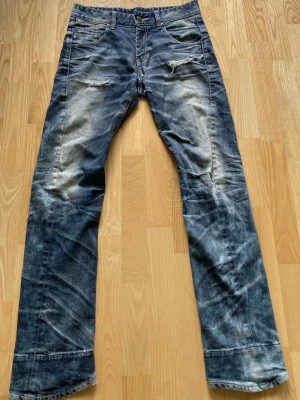 Evisu byxor  - Ett par blå jeans med slitningar och trasiga detaljer framtill, samt broderade detaljer och patch på bakfickorna. Jeansen har raka ben och tydliga slitningar vid benslut och lår. Klassisk femficksmodell i denim.