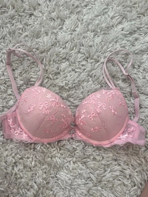 Rosa push-up bh med spets och rosetter - Söt rosa push-up bh med vadderade kupor, dekorerad med broderade rosetter och blommor i spets. Justerbara axelband och liten rosett med strassdetalj framtill. Perfekt för dig som gillar romantiska detaljer och mjuka pastellfärger.