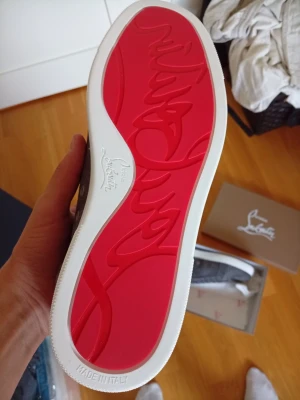 Gråa sneakers med röd sula - Snygga gråa sneakers i mocka från Christian Louboutin med den ikoniska röda sulan och vita kanter. Skorna har klassisk låg profil, snörning och en stilren design som sticker ut. Perfekta för dig som vill ha något exklusivt och trendigt.