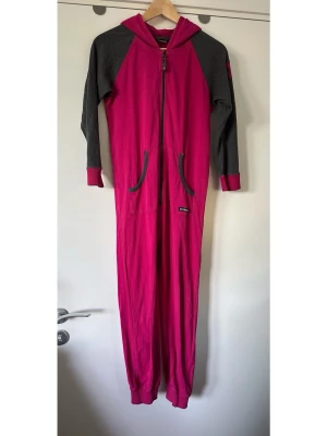 Villervalla - rosa jumpsuit / overall med huva - storlek 140 - Fin rosa overall / one-piece från Villervalla i storlek 140 med huva och dragkedja framtill. Långa ärmar i grå kontrastfärg med muddar vid handlederna och långa ben med muddar vid anklarna. Två fickor framtill. Mjukvävt tyg i 100% bomull, så bra naturmaterial som andas. Mycket gott skick!