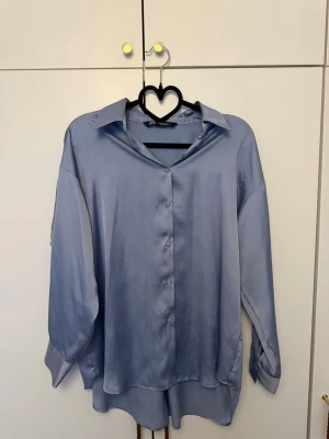 Ljusblå satinblus från Zara - Säljer en ljusblå skjorta från Zara i glansigt satinliknande material. Skjortan har klassisk krage, knappar framtill och långa ärmar med breda manschetter. Perfekt för en trendig och avslappnad look.