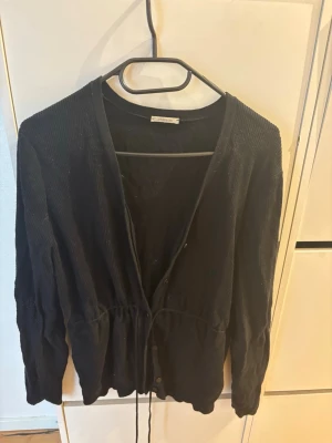 Svart ribbad kofta med knytband Intimissimi - Säljer en svart långärmad kofta med ribbad struktur och knytband i midjan. Koftan har knappar framtill och en avslappnad passform som gör den enkel att styla till olika looks. Perfekt för lager på lager och passar till det mesta.