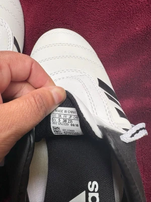 Vita Adidas fotbollsskor med svarta detaljer - Snygga vita fotbollsskor från Adidas med klassiska svarta ränder och svart häl. Skorna har snörning och är tillverkade i slitstarkt syntetmaterial. Perfekta för dig som vill ha stilrena och bekväma skor på planen.