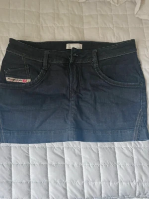 Mörkblå jeanskjol från Diesel - Snygg mörkblå jeanskjol från Diesel med klassiska fickor fram och bak, bälteshällor och dragkedja. Kjolen har en rak passform och är perfekt för dig som gillar en enkel men cool denimlook.