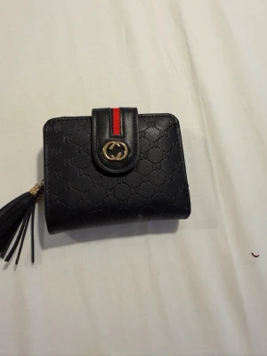 Svart plånbok med tofs och röd detalj - Snygg svart plånbok i konstläder med präglat mönster, guldfärgad logga och röd rand på knäppningen. Plånboken har dragkedjefack, flera fack för kort och en dekorativ tofs. Perfekt storlek för att få plats med det viktigaste.