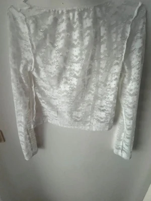Vit spetsblus med lång ärm - Säljer en vit, långärmad blus i spets med omlottdetalj framtill. Toppen har en feminin och elegant look med transparent spetsmönster och figurnära passform. Perfekt för dig som vill ha en romantisk vibe till din outfit.