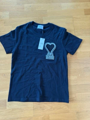 Svart t-shirt från Ami med hjärtdetalj - Snygg svart t-shirt från Ami med ett stort broderat hjärta och bokstaven A på bröstet. Klassisk rund halsringning och korta ärmar. Tillverkad i mjuk bomull som känns skön mot huden. Perfekt för en clean och trendig look.