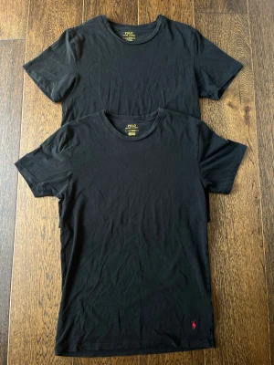 2 st t-shirts från Polo Ralph Lauren - Två stilrena svarta t-shirts från Polo Ralph Lauren med klassisk passform och rund hals. De har en liten röd logga broderad längst ner. Perfekta till jeans eller shorts för en enkel och snygg look.