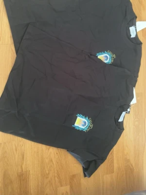 Svart t-shirt med tryck - Säljer en svart t-shirt med ett litet färgglatt tryck på bröstet. T-shirten har rund hals och korta ärmar, perfekt för en avslappnad stil. Materialet känns mjukt och bekvämt, troligen bomull.