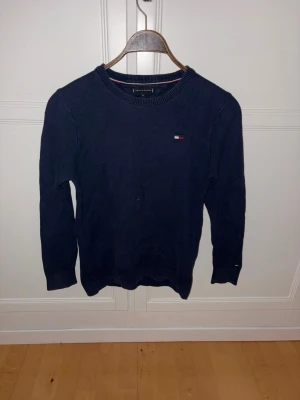 Mörkblå stickad tröja Tommy Hilfiger - Mörkblå stickad tröja från Tommy Hilfiger med rund halsringning och diskret logga på bröstet. Tröjan har långa ärmar och ribbade muddar vid ärmslut och nederkant. Perfekt för en stilren och enkel look. Passar perfekt för dig med storlek 164-176
