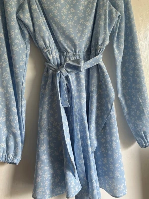 Ljusblå blommig omlottklänning - Söt ljusblå klänning med vita blommor. Knytband i midjan och resår i ryggen. Går att ha ärmarna upp men även off-shoulder. Volang detaljer i kjolen på klänningen. 