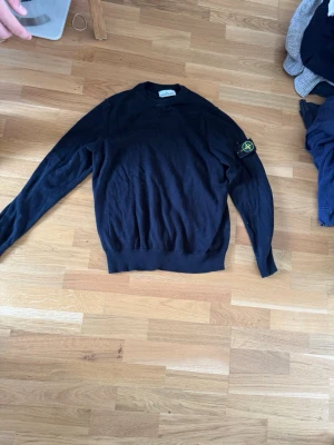 Svart stickad tröja Stone Island - Svart stickad tröja från Stone Island med klassisk rund hals och ribbade muddar. Tröjan har den ikoniska Stone Island-loggan på vänster ärm. Perfekt för dig som gillar streetwear och stilren design.  Vet ej om äkta