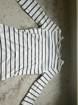 Randig långärmad topp från KappAhl - Vit långärmad topp från KappAhl med marinblå horisontella ränder. Toppen har en rund halsringning och är tillverkad i ett mjukt ribbat material som ger en skön passform. Perfekt för en avslappnad och stilren look.