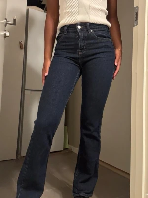 Mörkblå bootcut jeans med hög midja - Snygga mörkblå bootcut jeans med hög midja och klassiska fem fickor. Jeansen har vita kontrastsömmar och stängs med knapp och dragkedja. Materialet är denim med lätt stretch för skön passform. Perfekta för en trendig och avslappnad look.