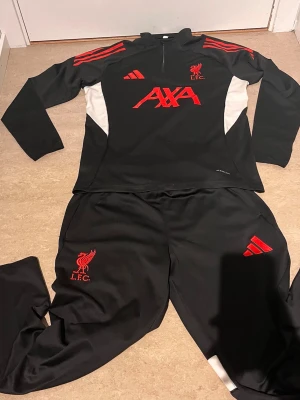 Liverpool FC träningsset Adidas svart/röd - Säljer ett svart träningsset från Adidas och Liverpool FC med röda detaljer och loggor. Setet består av långärmad tröja med halv dragkedja och matchande byxor. Materialet är mjukt och stretchigt, perfekt för träning eller uppvärmning. AXA-logga och LFC-märke på bröstet.