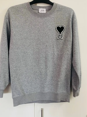 Grå sweatshirt från Ami Paris - Grå sweatshirt från Ami Paris med svart hjärta och A-logga broderad på bröstet. Klassisk rund halsringning, ribbade muddar och avslappnad passform. Perfekt för en stilren och trendig look.