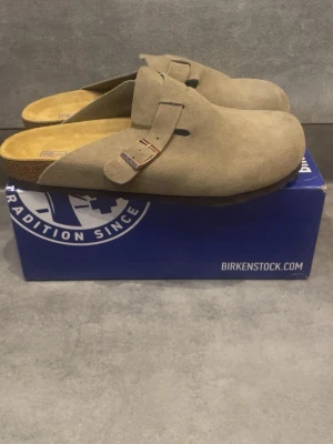 Birkenstock Boston taupe tofflor - Säljer ett par Birkenstock Boston tofflor i färgen taupe. De har en klassisk slip-in design med justerbart spänne och ovandel i mocka. Fotbädden är anatomiskt utformad och sulan är i kork och gummi för extra komfort. Perfekta för en chill och avslappnad stil.