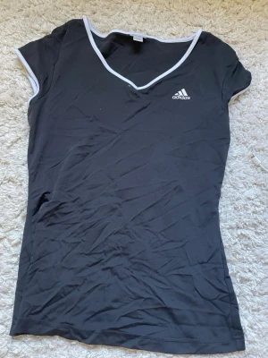 Svart v-ringad topp från Adidas - Svart topp från Adidas med vit kant runt v-ringningen och ärmsluten. Kortärmad och figurnära modell med diskret Adidas-logga på bröstet. Perfekt för en sportig och avslappnad stil.