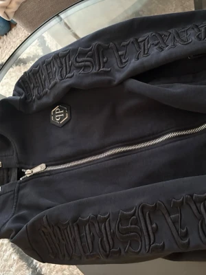  Philipp Plein hoodie  - Säljer en svart hoodie från Philipp Plein med dragkedja, stora broderade detaljer på ärmar och rygg samt en döskalle med korslagda ben på ryggen. Märkets logga i metall på bröstet. Hoodien har en huva och är gjord i mjukt bomullsmaterial.
