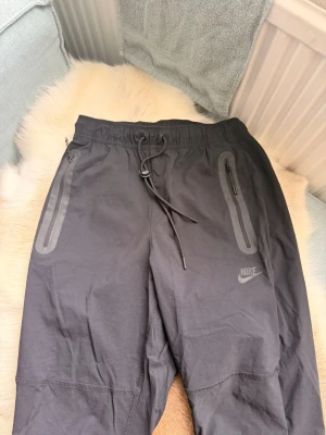 Nya nike tech woven - Svarta mjukisbyxor från Nike med elastisk midja och dragsko. Byxorna har två fickor med dragkedja och diskret Nike-logga på vänster ben. Tillverkade i ett mjukt och lätt material, perfekta för en avslappnad stil. Baggy passat dig som har M