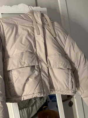 Beige pufferjacka med huva - Snygg beige pufferjacka med stor huva och två stora fickor framtill med lock och tryckknappar. Jackan har dragsko i huvan och är vadderad för extra värme. Perfekt för kalla dagar och enkel att matcha med olika outfits. stroeek S 