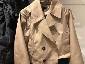Kort beige trenchcoat från Zara - Trendig kort trenchcoat från Zara i beige bomullsmix. Jackan har bred krage, dubbelknäppning med svarta knappar och axelklaffar. Två snedställda fickor framtill och markerade sömmar ger en cool look. Perfekt för lager på lager och streetstyle.