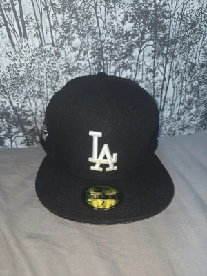 Svart LA Dodgers keps New Era - Säljer en svart LA Dodgers keps från New Era med broderad vit LA-logga framtill och World Series-märke på sidan. Modellen är 59FIFTY med platt skärm och klassisk passform. Perfekt för dig som gillar streetwear och sportig stil.