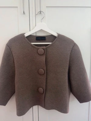 Beige kofta med stora knappar - Stilren beige kofta från Zara med tre stora klädda knappar framtill. Koftan har rund halsringning, rak passform och trekvartsärmar. Materialet är tjockstickat och ger en exklusiv känsla. Perfekt för dig som gillar minimalistisk och trendig stil.