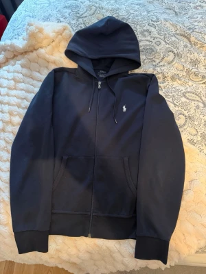  Polo Ralph Lauren zip hoddie - Klassisk marin blå zip hoodie från Polo Ralph Lauren med dragkedja framtill, två fickor och vit broderad logga på bröstet. Huva med dragsko och ribbade muddar. Perfekt för en chill och stilren look. Otroligt bra skick!