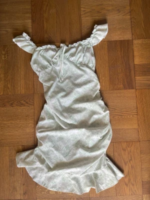 Ljus Blommig Off Shoulder Klänning med Volanger - Vit off shoulder klänning med grönt blommigt mönster. Volanger på axlarna och den nedre kanten. Rynkad i sidorna. Knytning vid bysten. Finns två små beiga fläckar, se de sista två bilderna. Finns ingen etikett så storleken är en uppskattning.