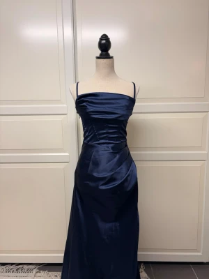 Marinblå långklänning i satin - Elegant marinblå långklänning med smala axelband och draperad överdel. Klänningen har en figurnära passform upptill och faller mjukt längs kroppen. Tillverkad i glansigt satinmaterial som ger en lyxig känsla. Vid frågor om mått eller annat är det bara att höra av sig💕