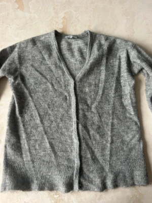 Ljusgrå stickad kofta - Mysig ljusgrå stickad kofta med v-ringning och långa ärmar. Koftan har en enkel och klassisk design utan knappar. Perfekt att slänga över en t-shirt för en avslappnad look.