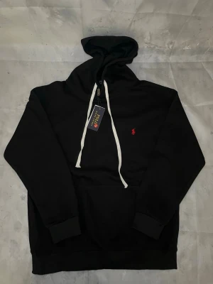 Svart hoodie från Polo Ralph Lauren - Snygg svart hoodie från Polo Ralph Lauren med vit dragsko i huvan och klassisk röd logga broderad på bröstet. Hoodien har känguruficka framtill och ribbade muddar vid ärmar och nederkant. Perfekt för en avslappnad och stilren look.