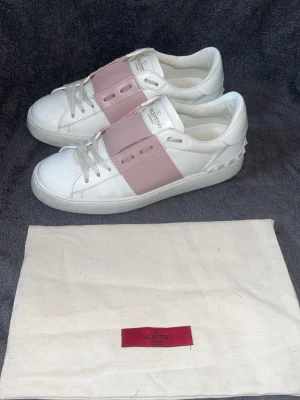 Valentino open - Snygga vita sneakers från Valentino med bred rosa läderdetalj över snörningen och ikoniska nitar på hälen. Skorna har en klassisk låg silhuett och är tillverkade i läder med vita snören och sula. Perfekta för dig som vill ha en exklusiv touch till din outfit.