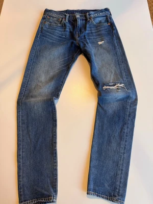 Levi's 512 blå slitna jeans - Säljer ett par klassiska blå Levi's 512 jeans med raka ben och snygga slitningar på låret och knät. Jeansen har fem fickor, gylf med dragkedja och den ikoniska läderlappen bak i midjan. Perfekta för en avslappnad och trendig look.