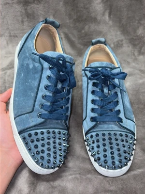 Washed blue sneakers med nitar från Louboutin - Unika blå sneakers i mocka från Christian Louboutin med matchande blå skosnören och coola metallnitar på tån. Skorna har vit sula och svarta detaljer längs sömmarna. Perfekt för dig som vill sticka ut med din stil.