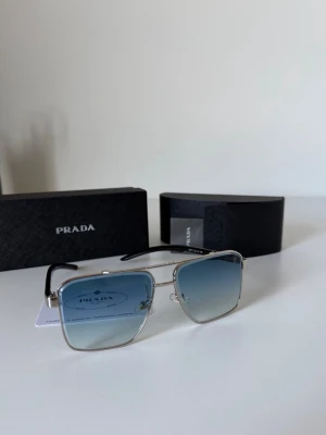 Prada fyrkantiga solglasögon silver - Snygga solglasögon från Prada med fyrkantiga bågar i silverfärgad metall och blåtonade glas. Skalmarna är svarta med en röd detalj och Prada-logga på sidan. Perfekt accessoar för dig som vill ha en modern och exklusiv look.