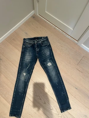 Mörkblå skinny jeans med slitningar - Säljer ett par mörkblå skinny jeans med coola slitningar och distressed detaljer på framsidan. Jeansen har fem fickor, knappgylf och smal passform. Perfekta för en avslappnad och trendig look.