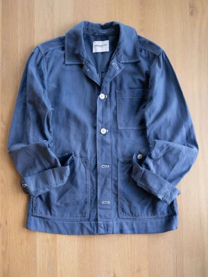 Blå overshirt / Chore Jacket från These Glory Days - Stilren blå overshirt från These Glory Days i bomull. Klassisk krage, tre utanpåliggande fickor och vita knappar framtill. Perfekt lager-på-lager-plagg med avslappnad passform och synliga sömmar för en modern look.