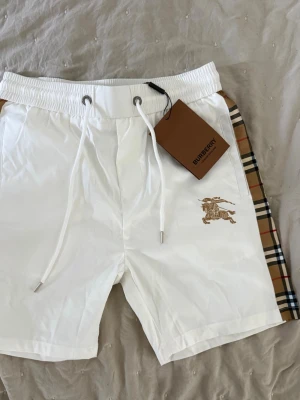 Vita Burberry shorts med rutiga sidor - Snygga vita shorts från Burberry med klassiska rutiga paneler på sidorna och broderad logga framtill. Storlek L men sitter som S. Elastisk midja med snörning för skön passform. Perfekta för en trendig och avslappnad look.
