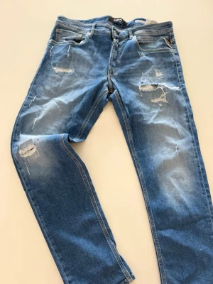Blå slitna jeans från Replay - Säljer ett par blå jeans från Replay med snygga slitningar och hål på benen för en trendig look. Jeansen har klassisk femficksmodell, normal midja och raka ben. Tillverkade i mjukt denimtyg som sitter skönt hela dagen.