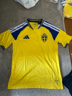 Sverige matchtröja Adidas gul/blå - Gul och blå matchtröja från Adidas med Sveriges landslagsemblem på bröstet. Tröjan har blå detaljer på axlarna, vita ränder och rund halsringning med blå och gula inslag. Tillverkad i lätt och ventilerande material, perfekt för fotboll eller träning.