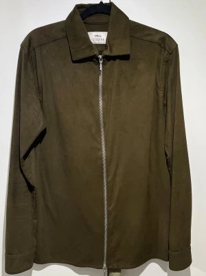 Olivgrön overshirt från Ciszere - Stilren olivgrön overshirt från Ciszere med rak passform och hel dragkedja framtill. Tillverkad i mjukt material som ger en modern look. Klassisk krage och långa ärmar gör den enkel att matcha med olika outfits.