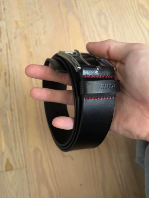 Svart skärp från Hugo Boss i skinn - svart bälte från Hugo Boss köpt för 600 aldrig använt i äkta skinn med silverfärgat spänne och röda sömmar som detalj. Perfekt accessoar för att lyfta din outfit med en stilren och modern touch. Märket syns diskret på lädret nära spännet.