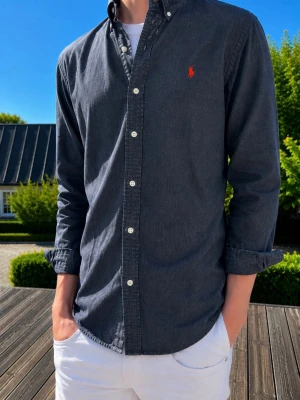 Mörkblå skjorta från Ralph Lauren - (MODDELEN ÄR 185 OCH VÄGER 75kg)Stilren mörkblå skjorta från Ralph Lauren med klassisk button-down krage och vit knappslå. Ikoniska röda loggan broderad på bröstet. Skjortan har långa ärmar och är tillverkad i ett mjukt bomullstyg. Perfekt för en avslappnad men snygg look.